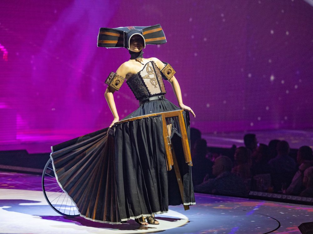 Model wearing black and brown gown with bicycle wheel behind onstage